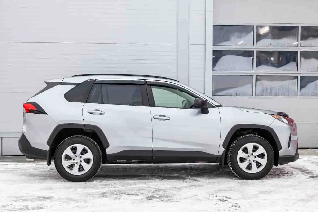 SUV Toyota RAV4 Toyota RAV4 blanc garé sur neige devant un bâtiment vitré, profil conducteur visible, version SUV avec finitions noires typiques des modèles récents.