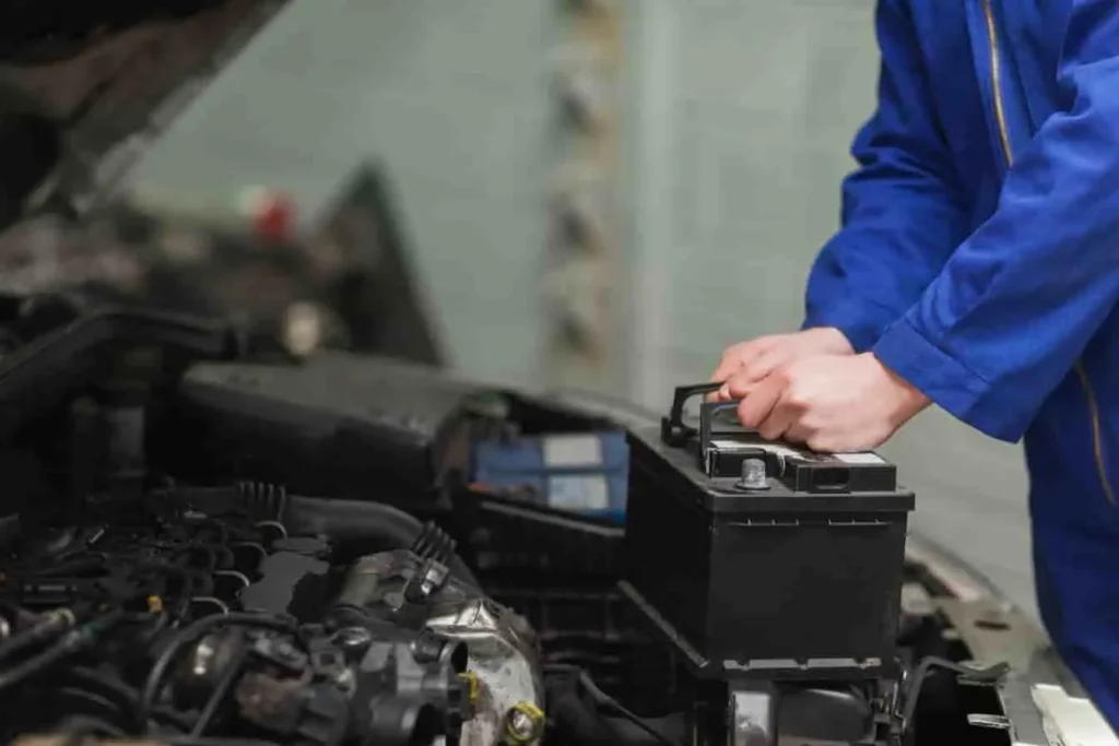 Mécanicien en bleu retirant une batterie de voiture sous le capot ouvert, entouré de composants moteur visibles.