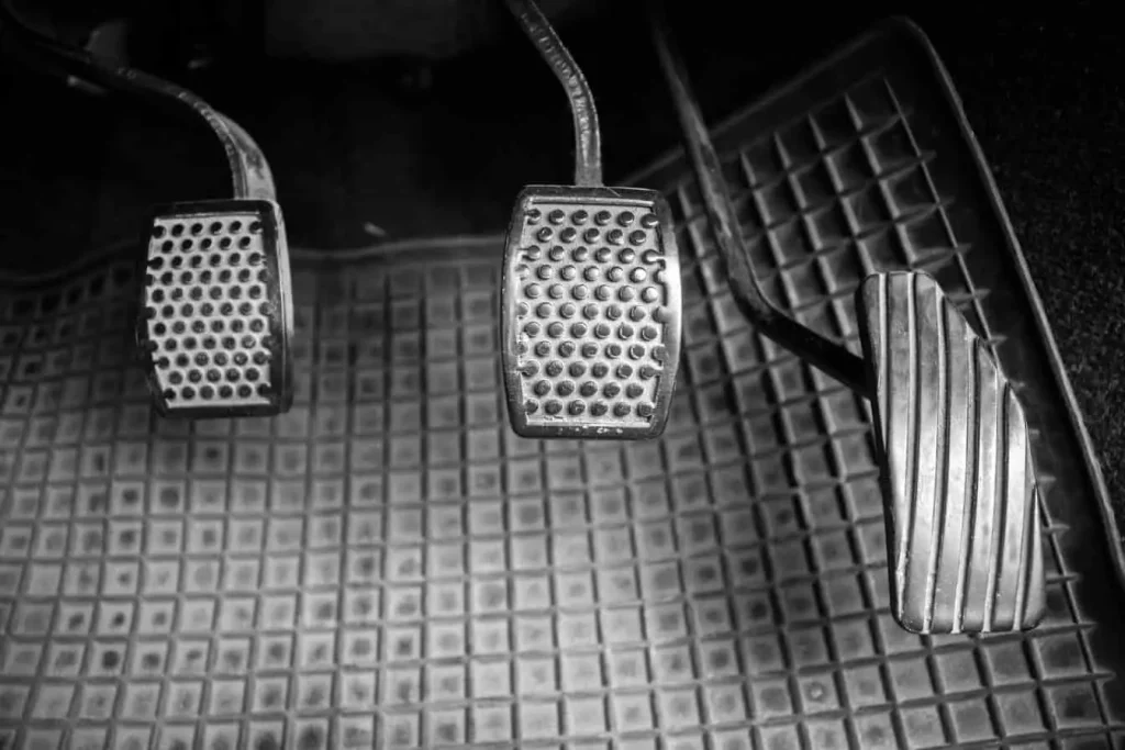 Photo en noir et blanc des pédales d'une voiture, embrayage, frein et accélérateur. Bruit métallique au freinage.
