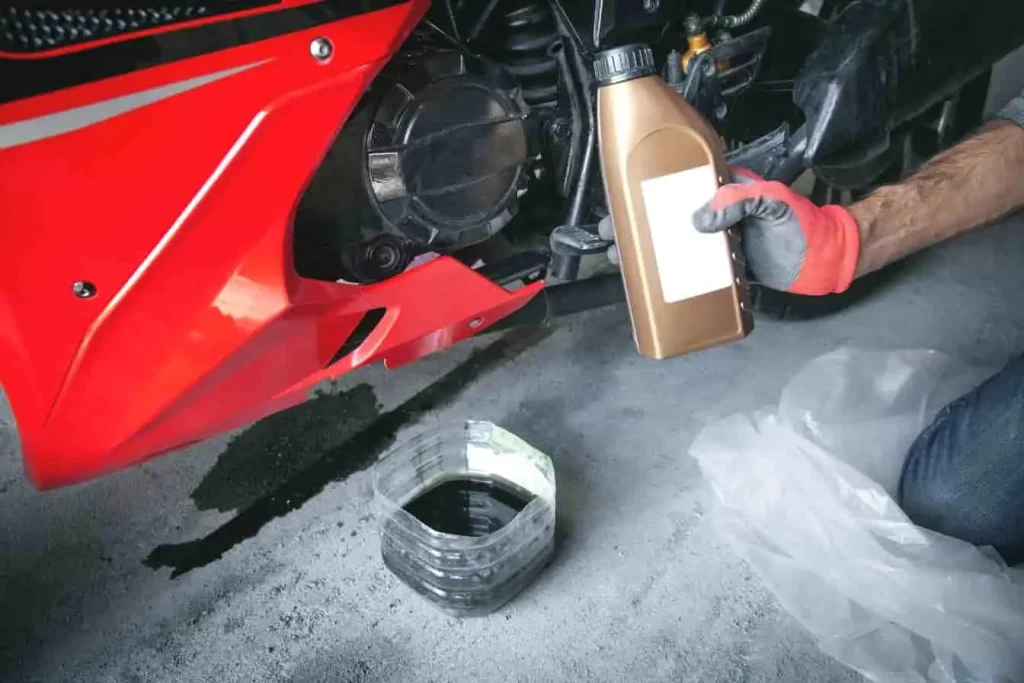 Vidange moto en cours : huile usagée récupérée dans un bac, nouveau bidon prêt à être versé, sol protégé par une bâche.
