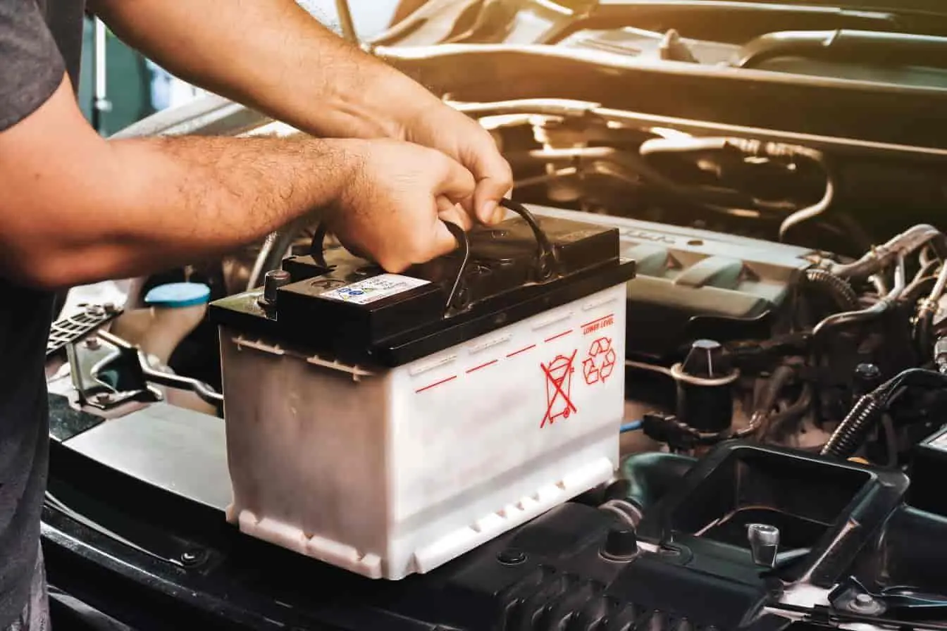 batterie de voiture Mains retirant une batterie de voiture dans le compartiment moteur, avec câbles visibles et étiquette de sécurité sur le boîtier.