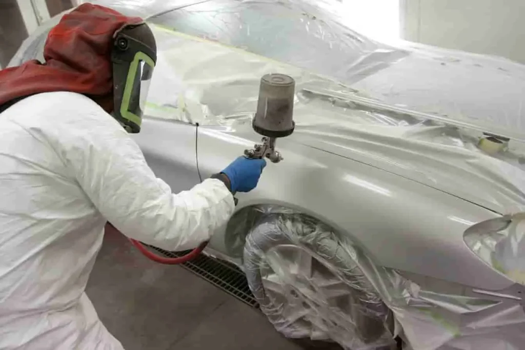 Carrosserie masquée avec plastique et ruban, professionnel en tenue intégrale pulvérisant une couche de peinture sur la surface