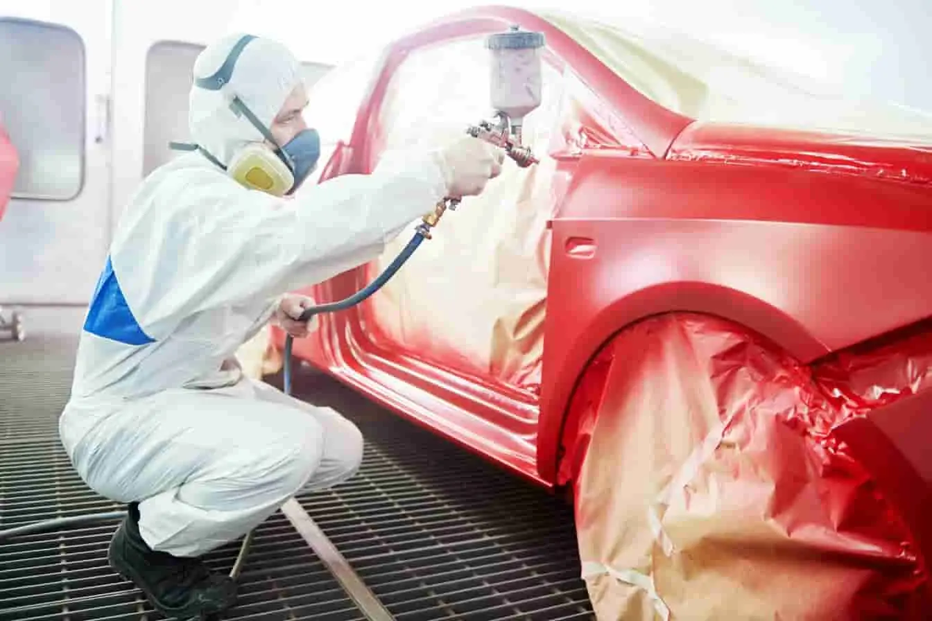 repeindre une voiture Carrosserie rouge en cours de peinture dans une cabine, professionnel en combinaison appliquant une couche uniforme avec pistolet