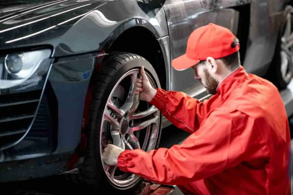 Mécanicien en uniforme rouge inspectant la roue avant d’un véhicule surélevé, vérification des causes possibles de vibrations au volant.
