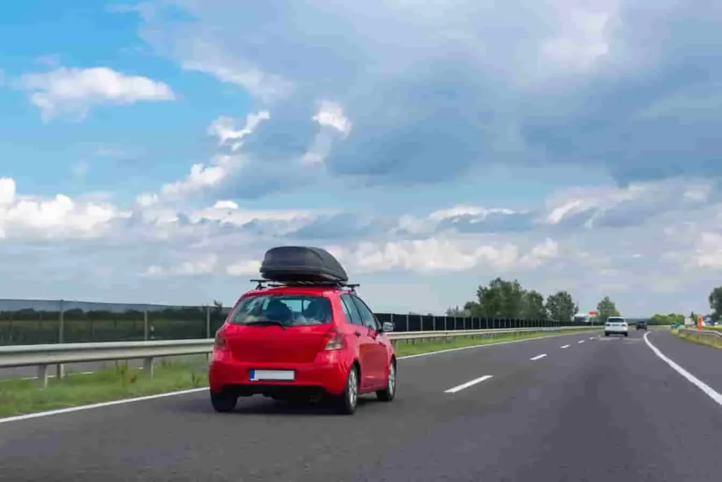 Voiture rouge roulant sur autoroute dégagée, équipée d’un coffre de toit noir, ciel partiellement nuageux