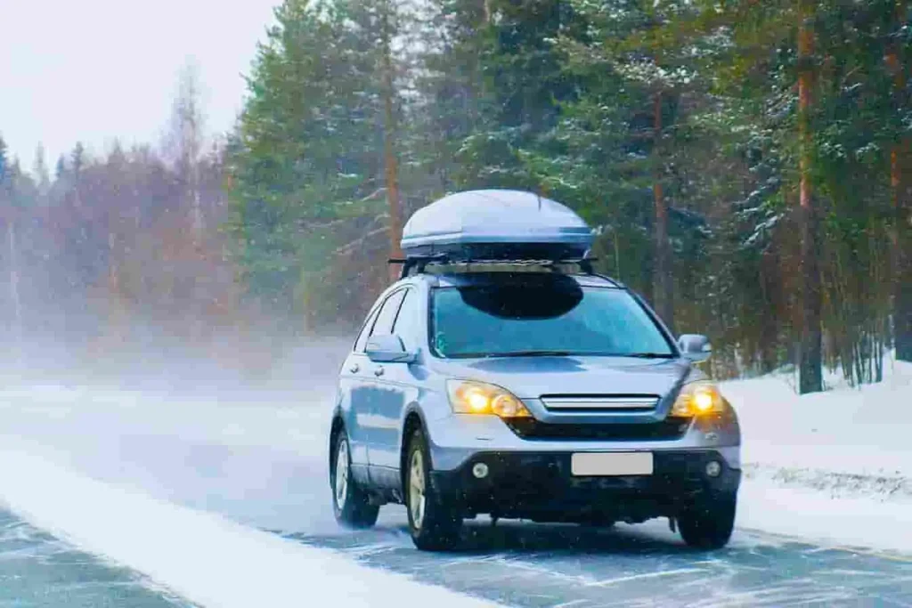 Voiture argentée avec coffre de toit traversant une route enneigée bordée d’arbres, neige soufflée par le vent