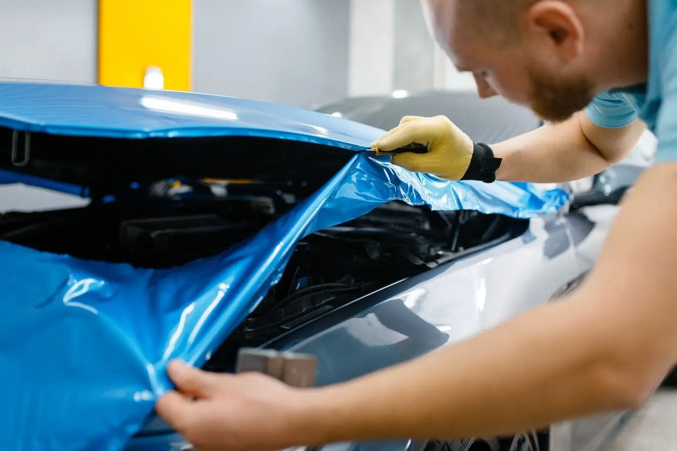 Main gantée appliquant un film bleu sur capot argenté, image d’un geste précis pour un covering durable