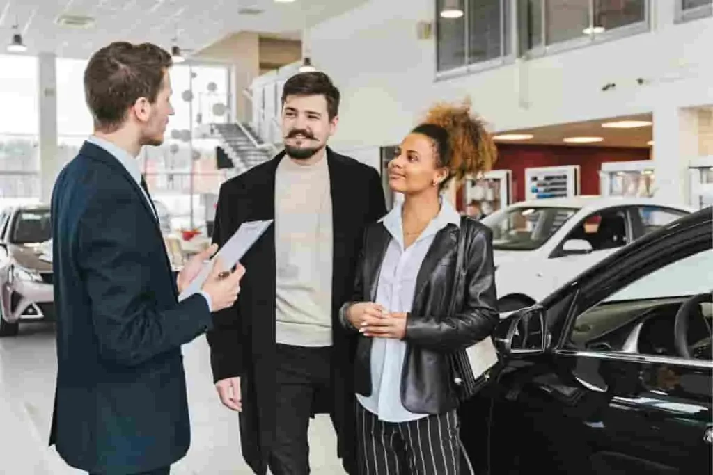 Un conseiller auto présente son offre à deux clients, illustrant le processus commercial en concession