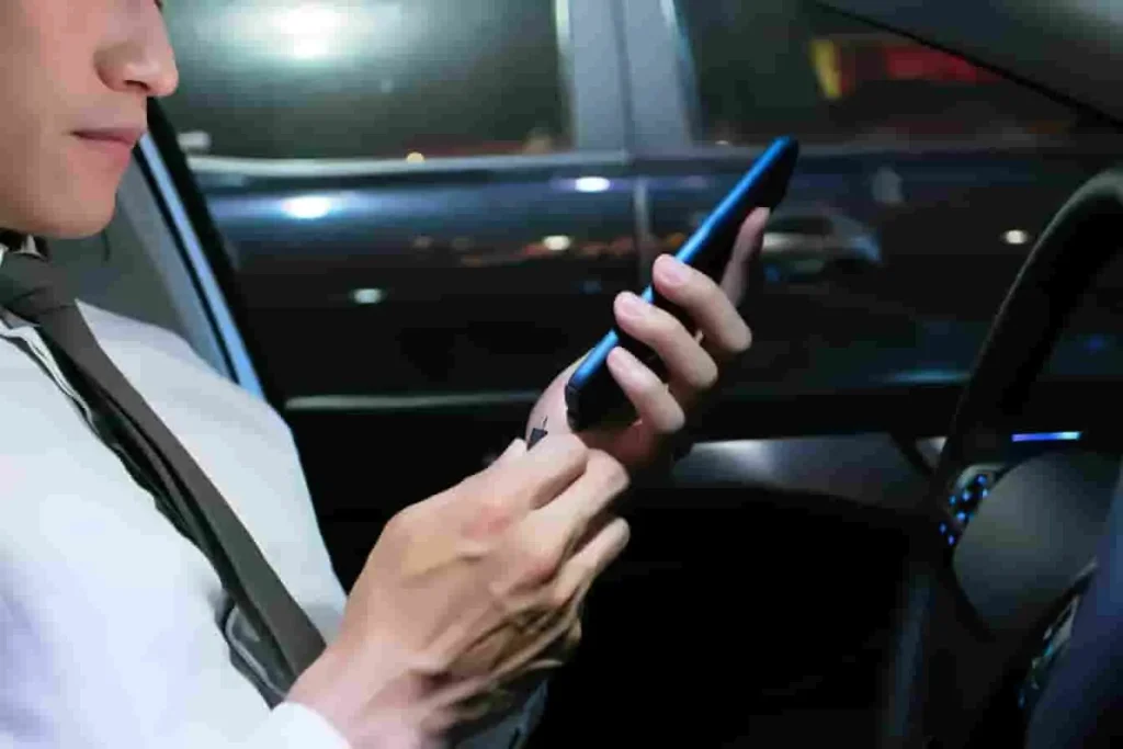 Personne en chemise blanche tenant un smartphone dans une voiture avec tableau de bord visible, image qui évoque une connexion Apple CarPlay en conduite
