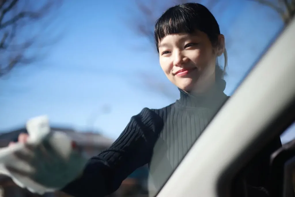Jeune femme utilisant un chiffon sur une vitre auto sous un ciel bleu pour montrer comment enlever de la colle sur une vitre de voiture sans acétone proprement