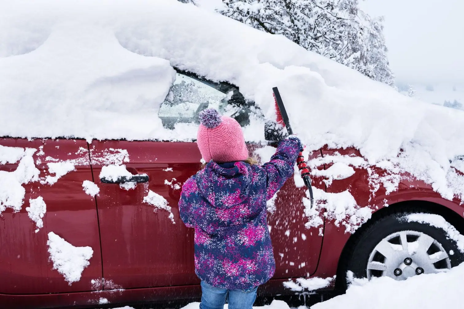 Voiture recouverte de neige Enfant dégage la neige d’une voiture rouge, geste préventif pour protéger la carrosserie du sel et éviter la corrosion hivernale