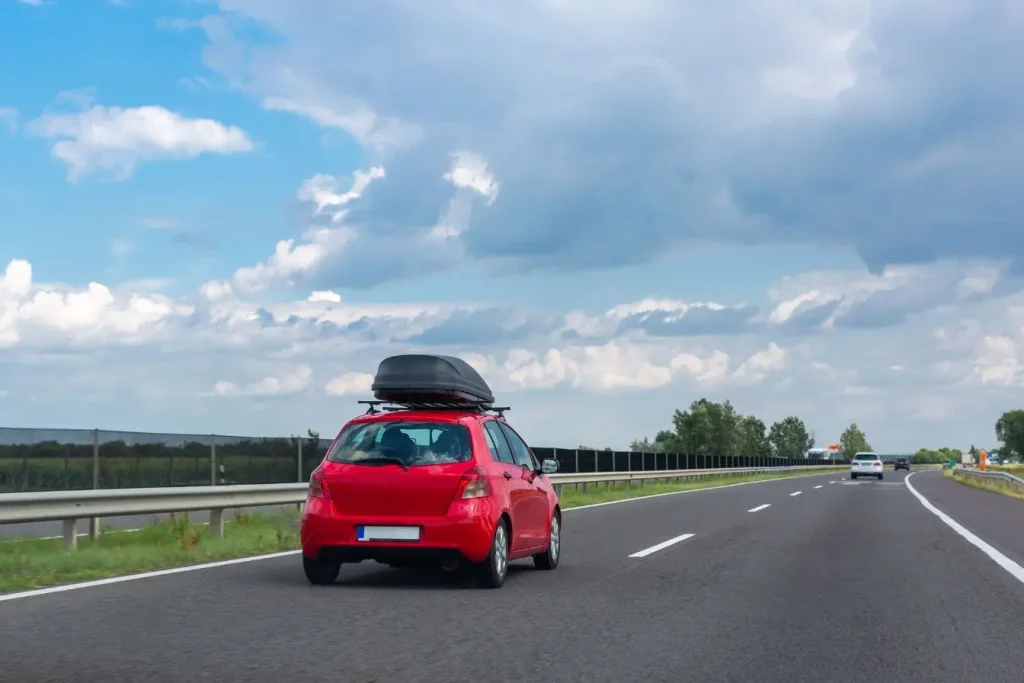 Voiture rouge roulant sur autoroute avec coffre de toit noir, image illustrant un accessoire pratique pour transporter des bagages en voyage