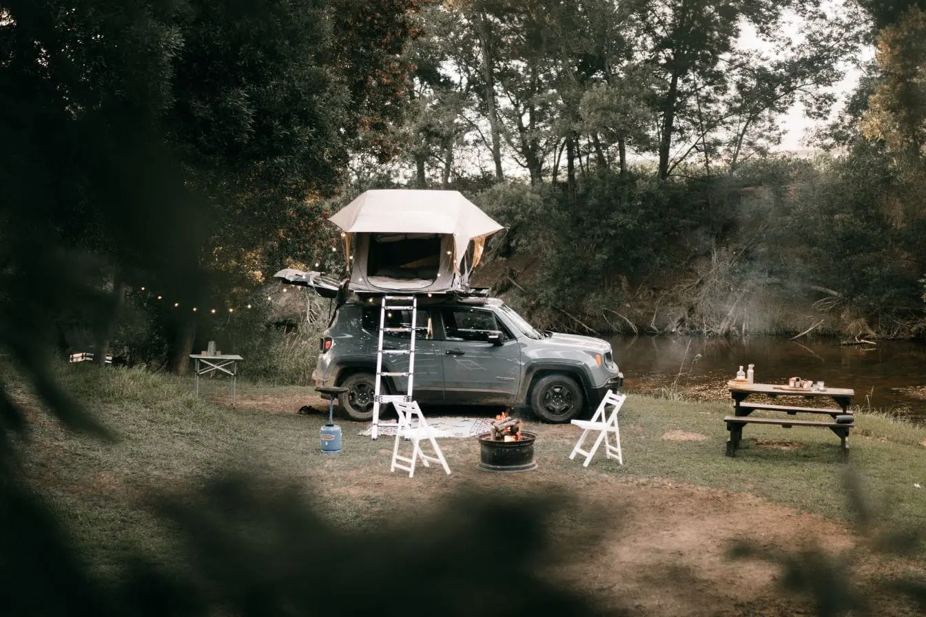 installer une tente de toit sur sa voiture SUV gris garé au bord d’une rivière avec tente de toit ouverte et échelle, illustrant l’art d’installer une tente de toit sur sa voiture pour camping.