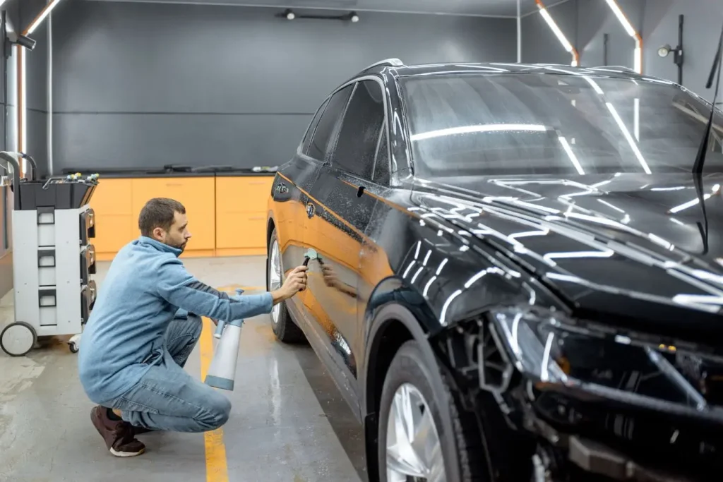 Personne accroupie utilisant spray et chiffon sur SUV noir brillant, illustrant l’application de cire de carrosserie pour protection et éclat