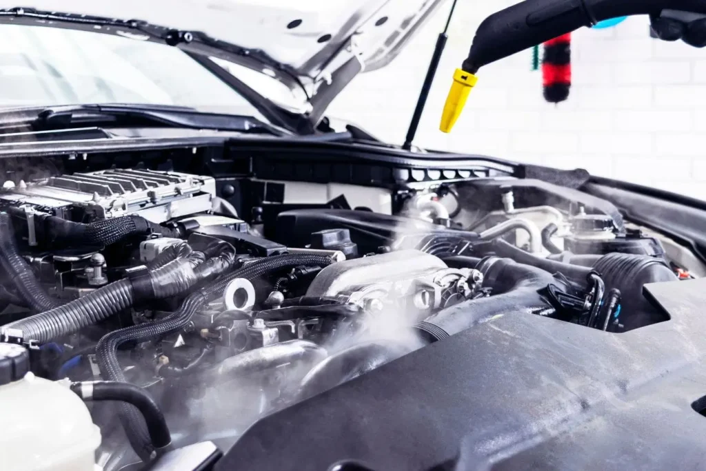 Compartiment moteur ouvert avec jet de vapeur dirigé sur pièces mécaniques, image illustrant l’action de nettoyer son moteur pour entretien automobile