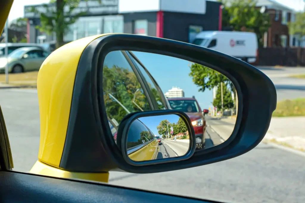 Rétroviseur d’une voiture jaune avec miroir additionnel reflétant route et véhicules derrière illustrant la vigilance et la détection d’angle mort.