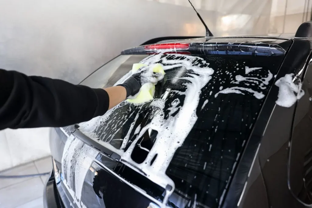 Main gantée nettoyant la vitre arrière d’une voiture noire avec éponge jaune et mousse, image illustrant l’usage du lave-glace pour une visibilité optimale