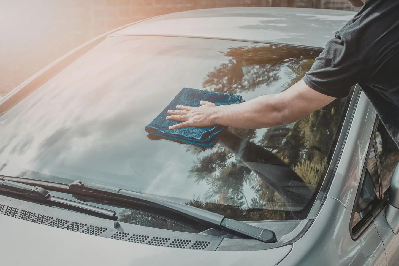 Nettoyage du pare-brise Personne nettoyant le pare-brise avec un chiffon bleu illustrant l’action d’enlever des autocollants sur une voiture pour retrouver une surface claire