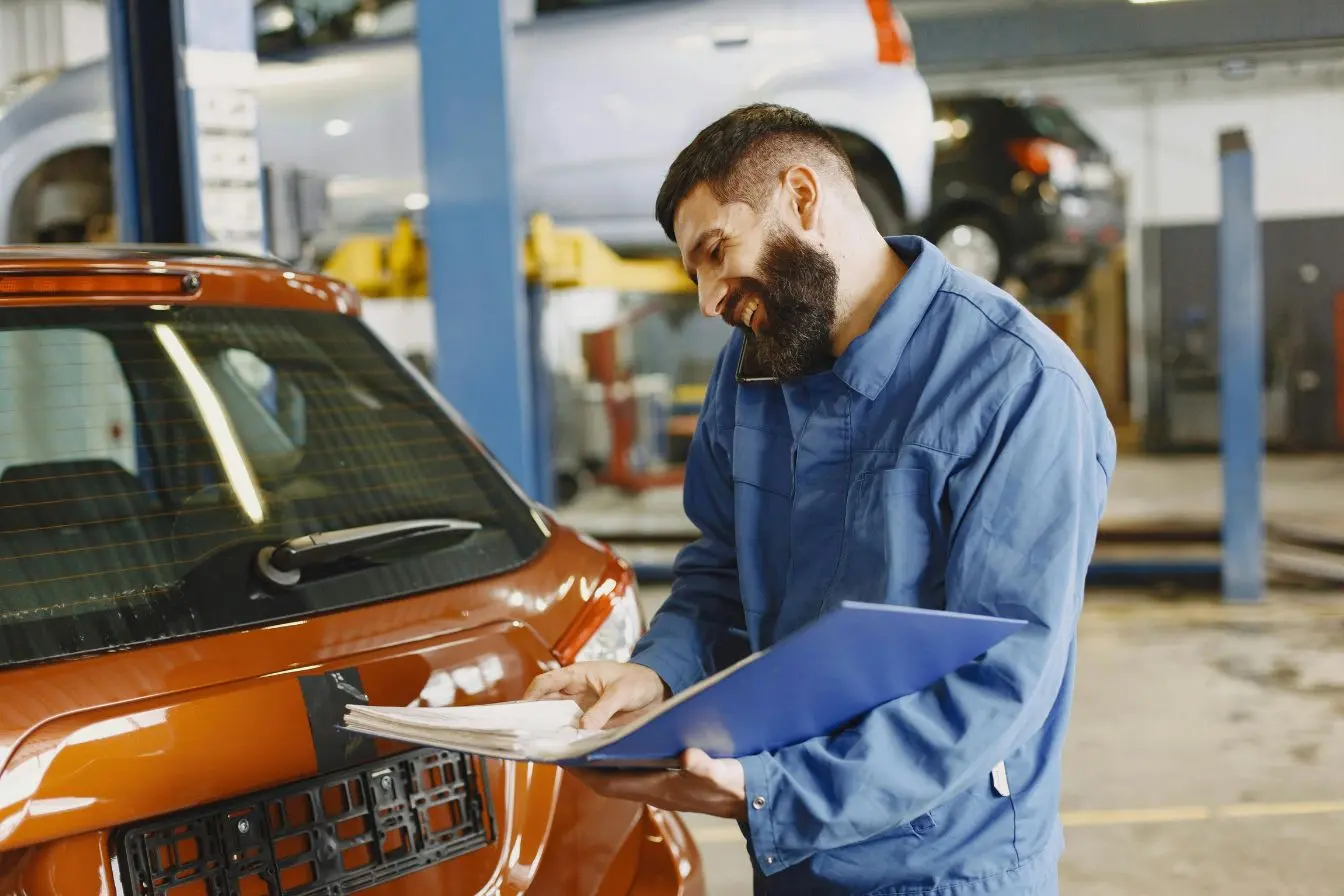 franchise en assurance auto Mécanicien en bleu de travail inspectant voiture orange dans garage, image illustrant la notion de franchise en assurance auto lors d’une réparation.