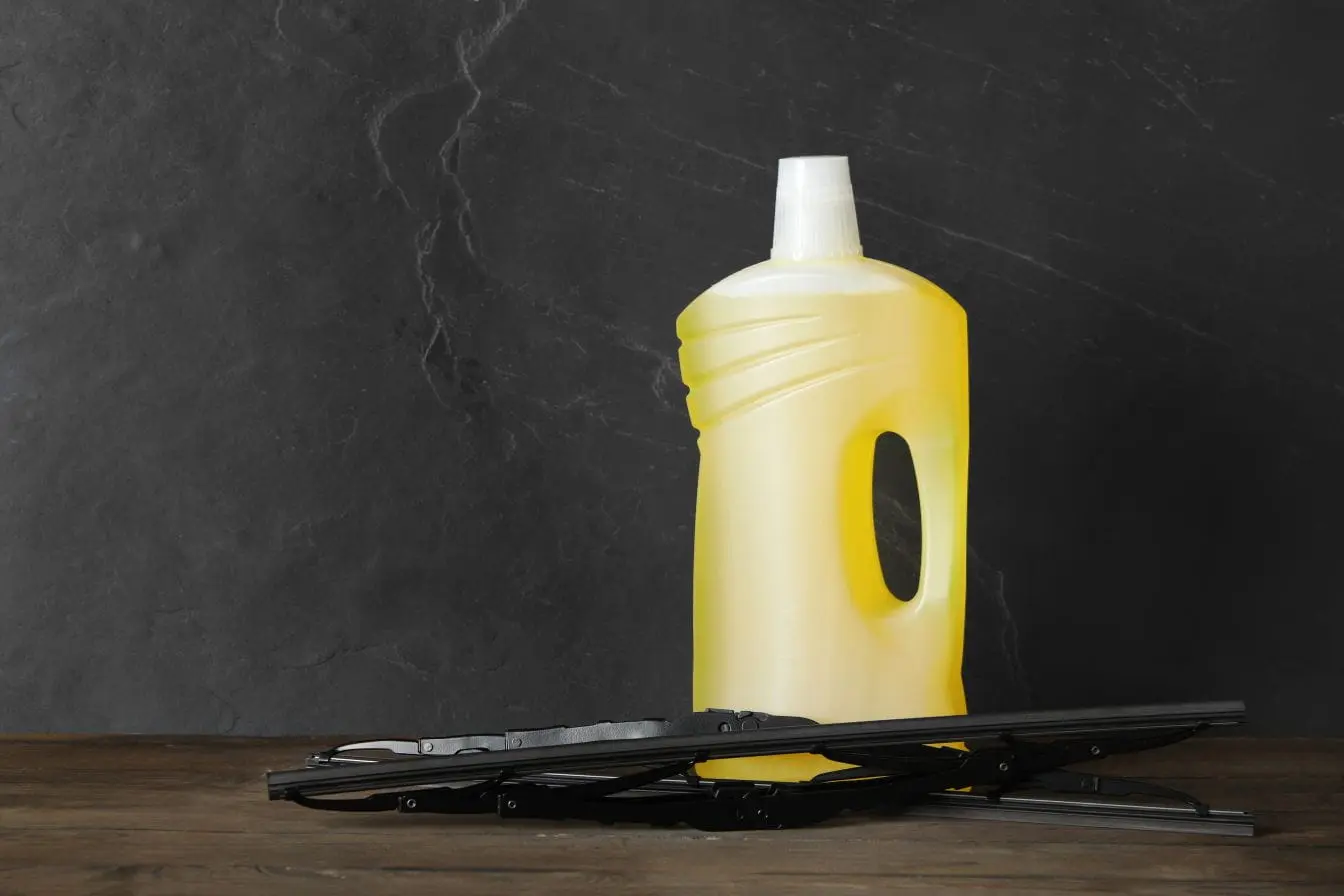 lave-glace Bidon jaune de liquide lave-glace posé sur table avec balais d’essuie-glace noirs, image illustrant l’entretien automobile