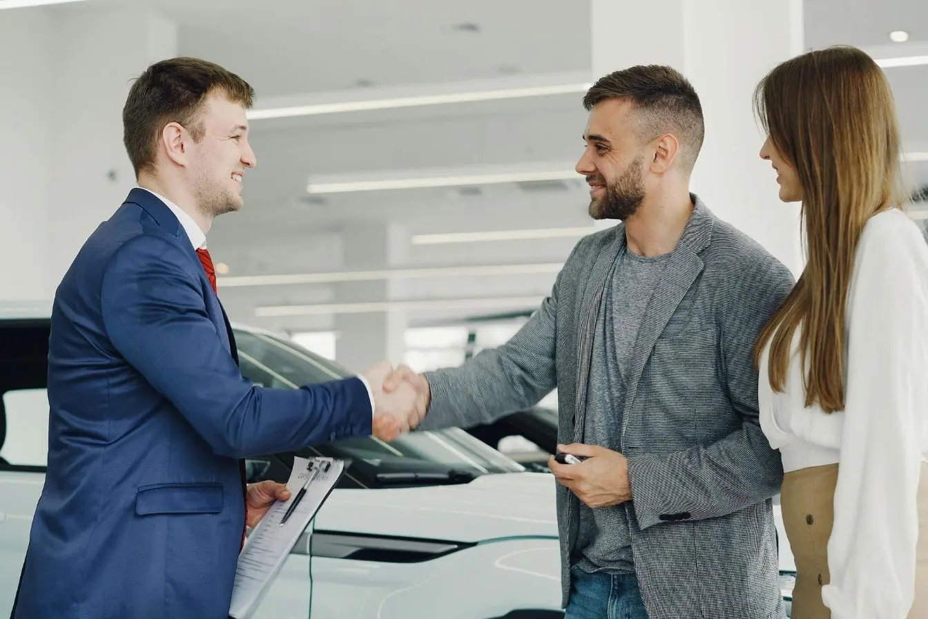 argus voiture Trois personnes en tenue professionnelle serrant main devant voiture blanche, image illustrant l’évaluation argus voiture lors d’une transaction.