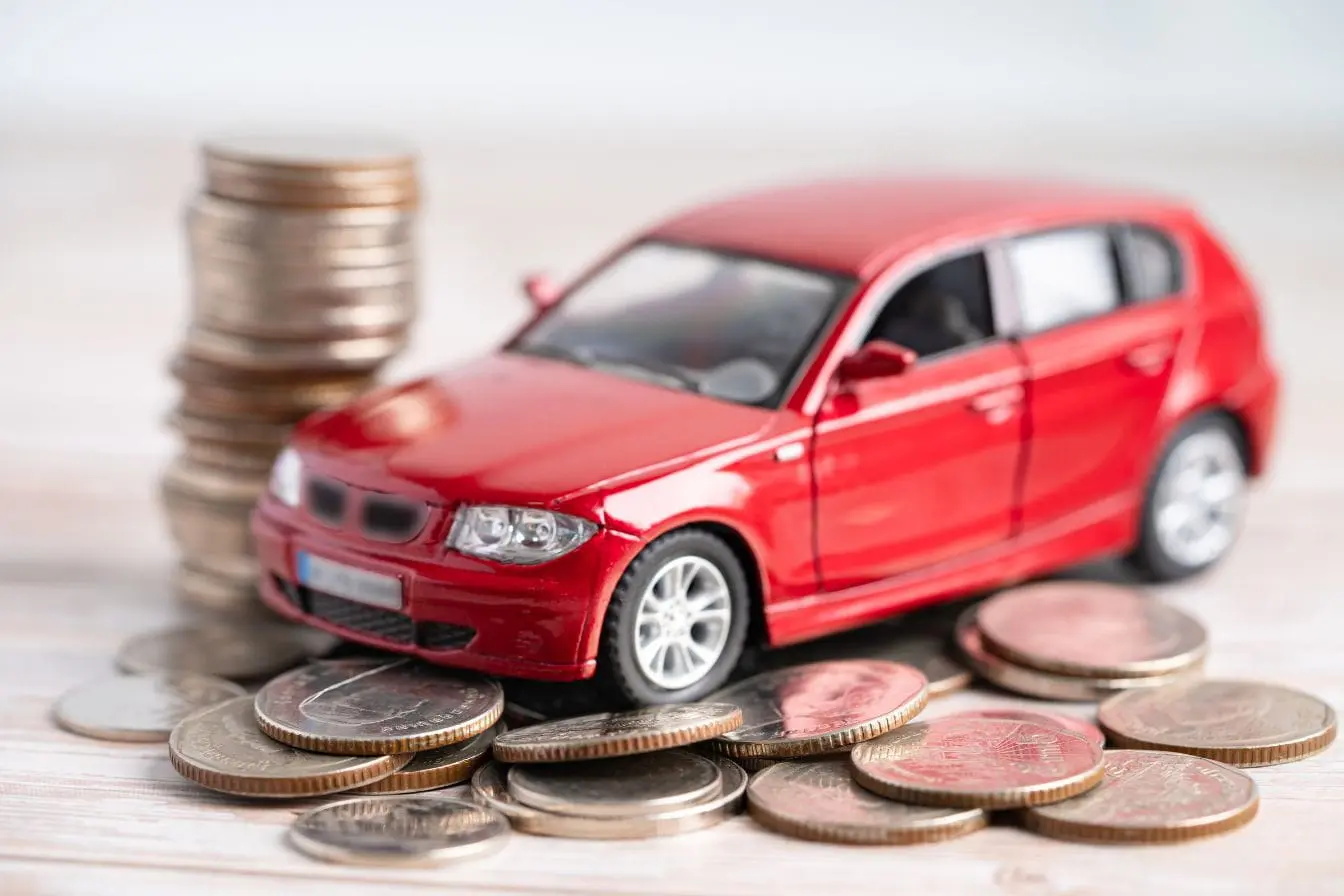 financement auto Petite voiture rouge entourée de pièces de monnaie illustrant le financement auto, entre épargne personnelle et coût d’acquisition.