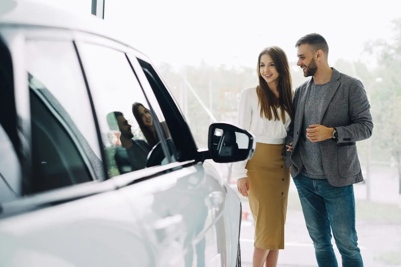 voiture neuve Deux personnes observant une voiture blanche en showroom, illustrant la décote immédiate d’une voiture neuve dès sa sortie du concessionnaire.