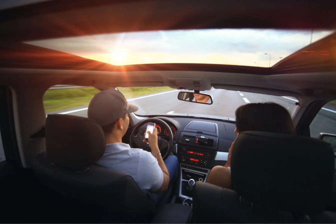 Vue intérieure d’une voiture Vue intérieure d’une voiture au coucher du soleil avec conducteur tenant un smartphone, image qui illustre l’importance du système de surveillance des angles morts.